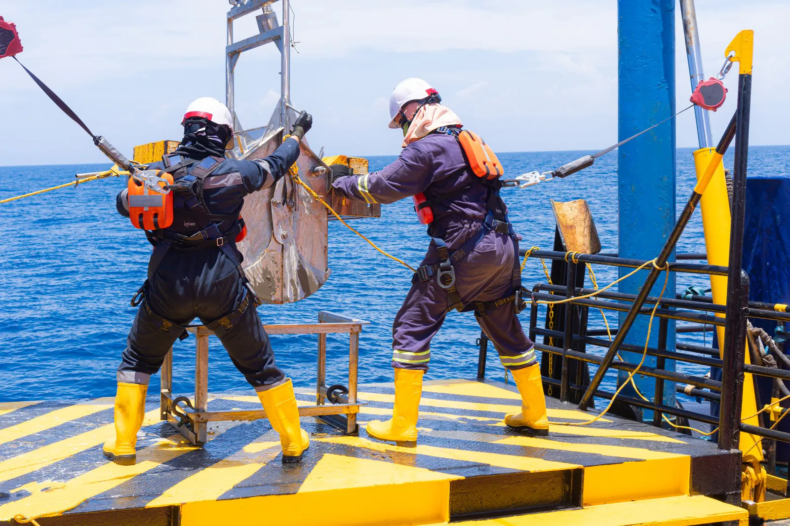 Two offshore workers in safety gear handling a suspended load on a platform deck
