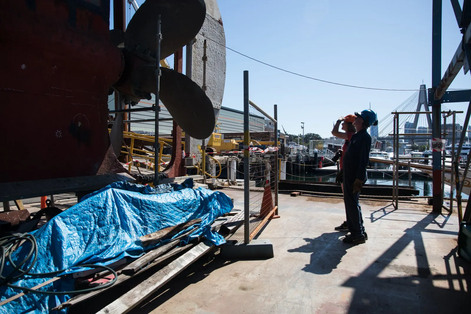 A ship propeller and hull in a dockyard with workers alongside